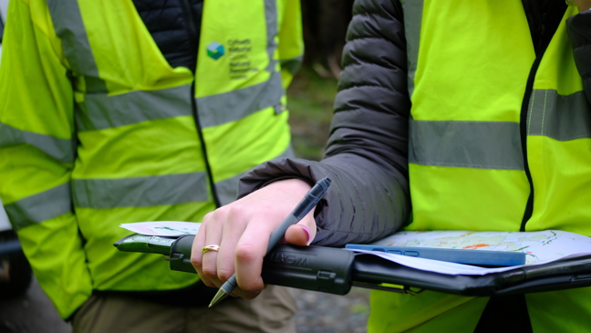 Two people stood, one is holding a clipboard. Both are in high-vis jackets. Faces are not shown.